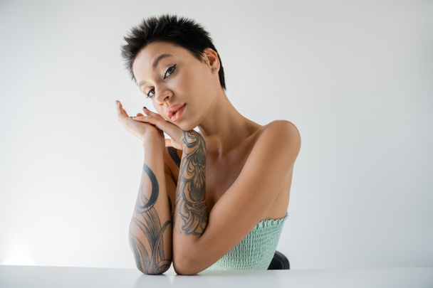 τατουάζ μελαχρινή γυναίκα σε στράπλες κορυφή κάθεται με τα χέρια κοντά στο πρόσωπο απομονώνονται σε γκρι - Φωτογραφία, εικόνα