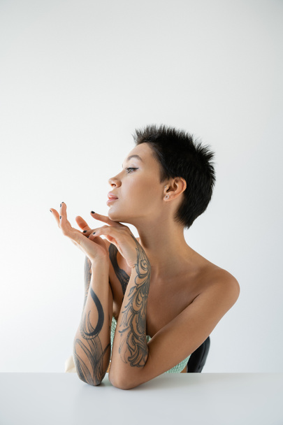 πορτρέτο της νεαρής γυναίκας με τατουάζ που κάθεται κοντά στο τραπέζι και κοιτάζει μακριά απομονωμένη στο γκρι - Φωτογραφία, εικόνα