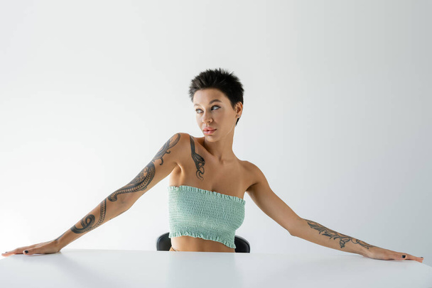 σέξι γυναίκα με τατουάζ σε στράπλες σουτιέν κάθεται στο τραπέζι και κοιτάζοντας μακριά σε γκρι φόντο - Φωτογραφία, εικόνα