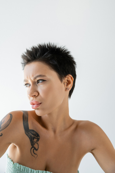 portrait of displeased tattooed woman in strapless top looking away isolated on grey - Foto, imagen