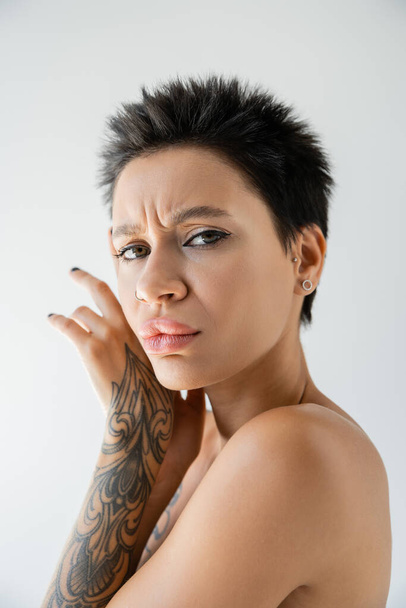 portrait of frowning woman holding tattooed hands near face and looking at camera isolated on grey - Photo, image