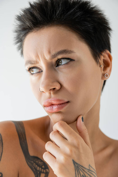 portrait of upset and frowning woman with makeup and piercing touching chin and looking away isolated on grey - Photo, Image