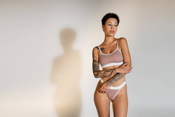 femme brune tatouée en lingerie regardant loin sur fond gris - Photo, image
