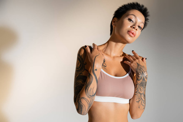 mujer morena tatuada con maquillaje posando en sujetador sobre fondo gris - Foto, imagen