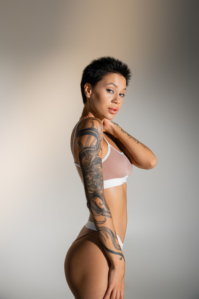 brunette woman with perfect tattooed body touching neck while posing in underwear on grey background - 写真・画像