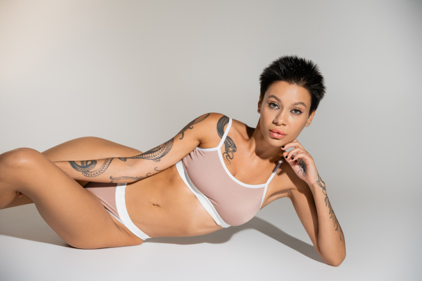 tattooed brunette woman in underwear looking at camera while posing on grey background - Photo, Image