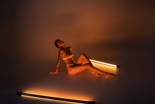 Полная длина соблазнительной женщины с тонким татуированным телом, сидящей в нижнем белье возле люминесцентных ламп на темном фоне - Фото, изображение