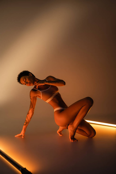 full length of sexy woman with perfect tattooed body posing in lingerie near fluorescent lamps on beige background - Valokuva, kuva