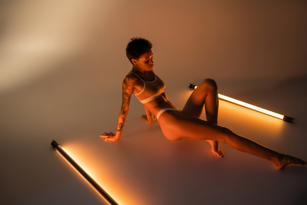 full length of brunette woman with tattooed body sitting in lingerie near fluorescent lamps on beige background - Photo, image