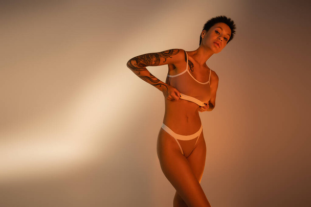 jovem mulher tatuada com corpo esbelto ajustando sutiã e olhando para a câmera no fundo bege - Foto, Imagem