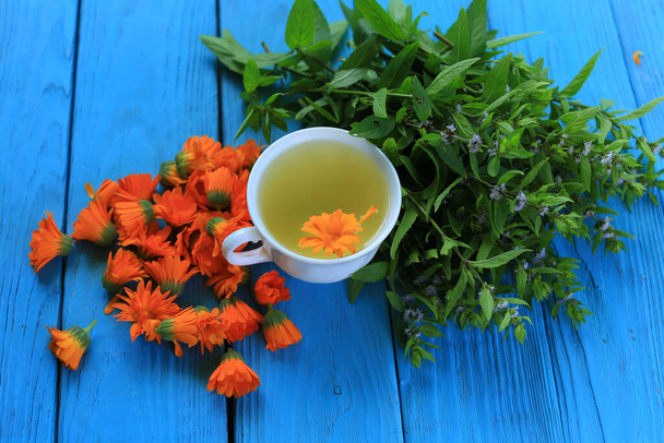 Biały porcelanowy kubek z herbata kwiat marigold na niebieskim drewnianym tle z bandą świeżych kwitnących mięty - Zdjęcie, obraz