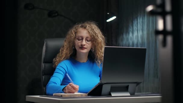 Curly woman learning online using laptop and notepad sitting at home.Young girl make notes with serious face in dark room.Lady doing education english online . High quality 4k footage - Záběry, video