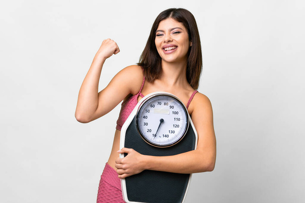 Young Russian woman over isolated white background with weighing machine and doing victory gesture - Fotó, kép