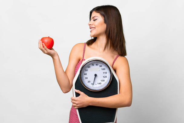 Young Russian woman over isolated white background holding a weighing machine while looking an apple - 写真・画像