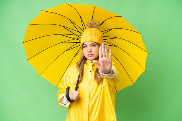 Jong meisje met regenbestendige jas en paraplu over geïsoleerde chroma key achtergrond maken stop gebaar - Foto, afbeelding