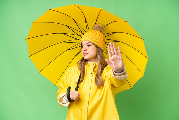 Menina com capa impermeável e guarda-chuva sobre fundo chave croma isolado fazendo parar gesto e decepcionado - Foto, Imagem