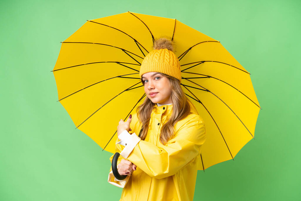 Chica joven con capa impermeable y paraguas sobre fondo de croma clave aislado apuntando hacia atrás - Foto, imagen