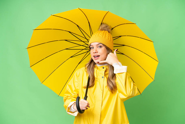 Jong meisje met regenbestendige jas en paraplu over geïsoleerde chroma belangrijkste achtergrond maken telefoon gebaar. Bel me terug. - Foto, afbeelding