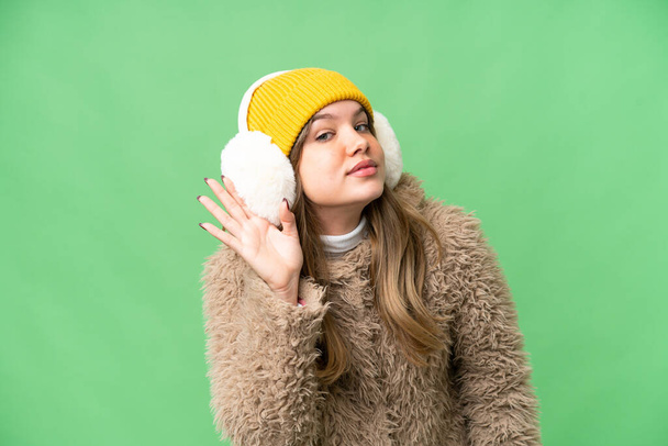 Νεαρό κορίτσι που φοράει μάσκες χειμώνα πάνω από απομονωμένο φόντο chroma κλειδί ακούγοντας κάτι βάζοντας το χέρι στο αυτί - Φωτογραφία, εικόνα