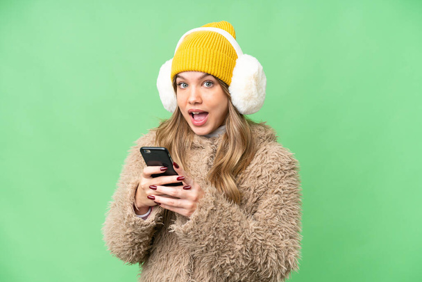 Chica joven con manguitos de invierno sobre fondo de croma clave aislado sorprendido y enviando un mensaje - Foto, Imagen
