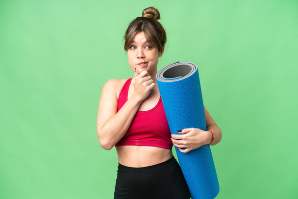 Young sport girl going to yoga classes while holding a mat over isolated chroma key background having doubts and thinking - Zdjęcie, obraz