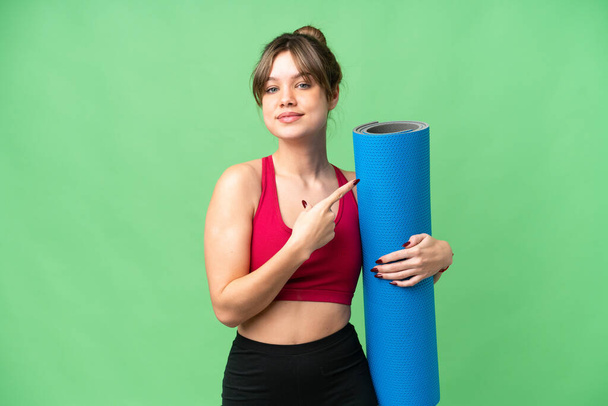 Junges Sportmädchen besucht Yoga-Kurse, während sie eine Matte über einen isolierten Chroma-Schlüsselhintergrund hält, der zur Seite zeigt, um ein Produkt zu präsentieren - Foto, Bild