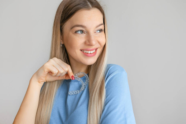 Beautiful smiling blonde woman is holding an invisible teeth aligner for orthodontic purposes. Includes copy space - Foto, imagen
