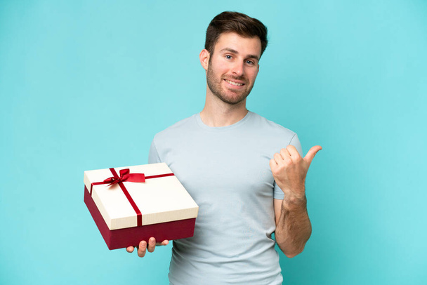 Young caucasian man holding a gift isolated on blue background pointing to the side to present a product - Photo, Image
