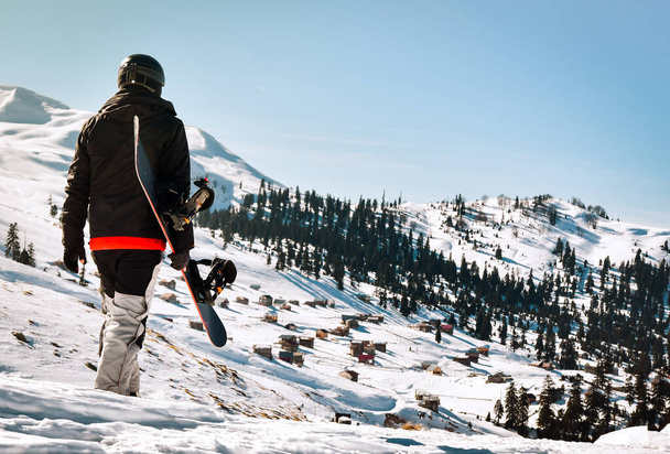 turista homem em traje de esqui com snowboard em suas mãos caminha na encosta coberta de neve contra o pano de fundo da bela paisagem montanhosa - Foto, Imagem