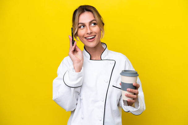 Uruguayan chef woman isolated on yellow background holding coffee to take away and a mobile - Φωτογραφία, εικόνα