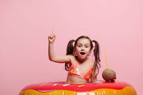 Αξιολάτρευτο καυκάσιο κοριτσάκι, που φοράει κομψό φωτεινό μαγιό, δείχνοντας με το δείκτη το χώρο διαφήμισης αντιγράφων, απομονωμένο σε ροζ φόντο, κοιτάζοντας την κάμερα, ποζάροντας με ποτό καρύδας και δαχτυλίδι κολύμβησης - Φωτογραφία, εικόνα