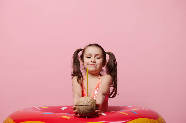 Γοητευτικό χαμογελαστό 5χρονο κοριτσάκι με μαγιό, που κρατάει στην κάμερα δροσιστικό coconut cocktail, ποζάρει με φουσκωτό donut swim ring σε ροζ φόντο. Πισίνα, καλοκαίρι, παραλία έννοια - Φωτογραφία, εικόνα