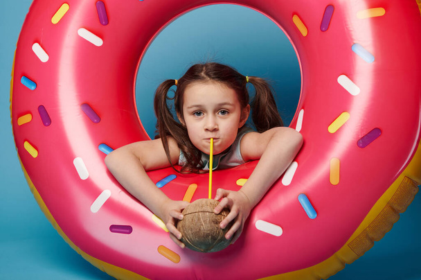 Caucasian adorable little girl in summer wear, drinking coconut water from a straw, posing with a pink inflatable swimming ring doughnut over blue color background. Tropical travel holidays concept - Zdjęcie, obraz