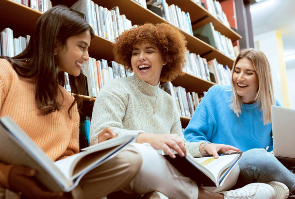Друзья, студенты в библиотеке с учебниками, университетская группа женщин, веселые и учащиеся вместе. Смех, разнообразие и молодость, исследование для бумаги или экзамена с мотивацией и чтением. - Фото, изображение