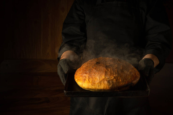 De bakker houdt vers gebakken warm brood in zijn handen op een pannetje in de bakkerij. Presentatie van bakkerijproducten. Plaats voor recept op donkere achtergrond - Foto, afbeelding
