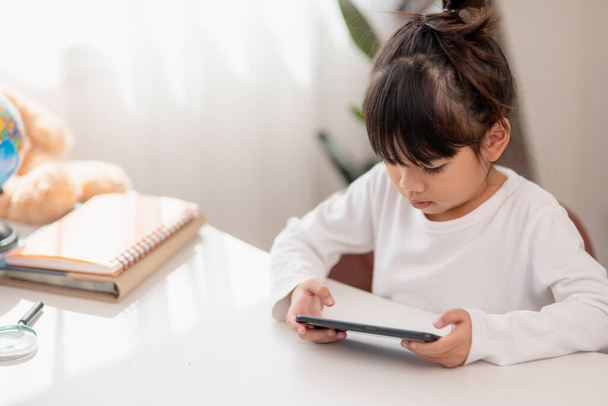 Ázsiai kislány használja a telefont közelről, eltereli a figyelmét a tanulásról, ül egy asztalnál jegyzetfüzetekkel, egy csinos gyerek szórakozik egy okostelefonnal, nézi a webináriumot, otthon tanul - Fotó, kép