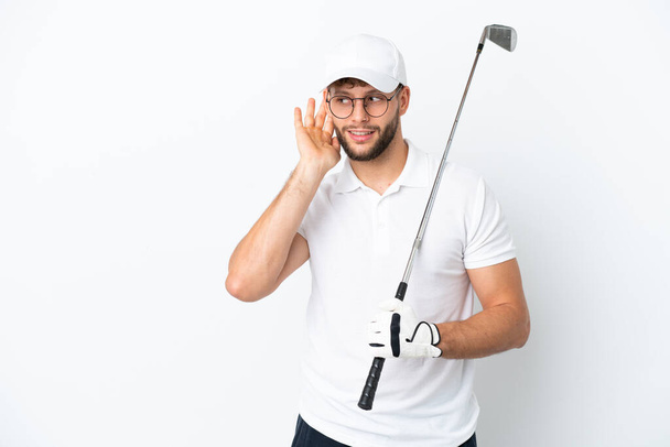 Bello giovane uomo che gioca a golf isolato su sfondo bianco ascoltando qualcosa mettendo la mano sull'orecchio - Foto, immagini