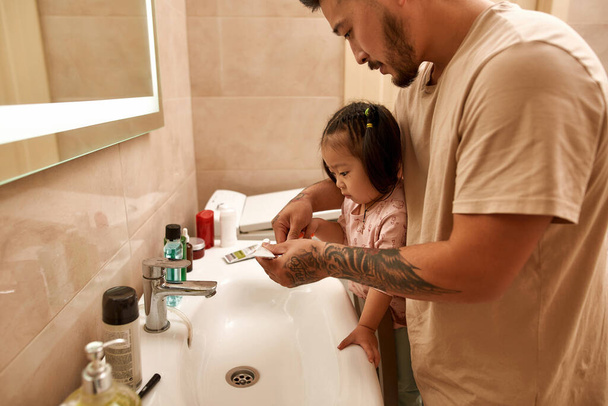 Side view of asian man pouring toothpaste on toothbrush of little daughter by wash basin in bathroom. Asian man and girl spending time together. Hygiene. Family relationship. Parenting and fatherhood - Photo, Image