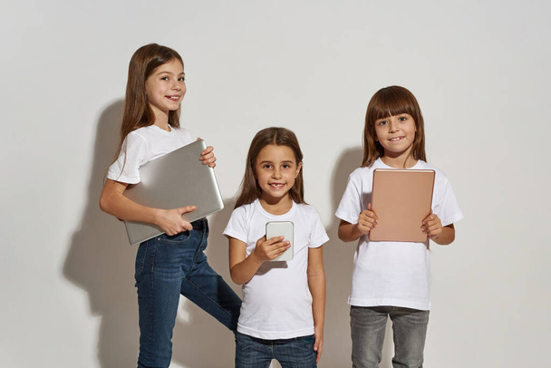 Smiling caucasian girls with variety digital gadgets. Female children with smartphone, digital tablet and laptop. Modern youngster lifestyle. Gadget addiction. White background in studio. Copy space - Zdjęcie, obraz