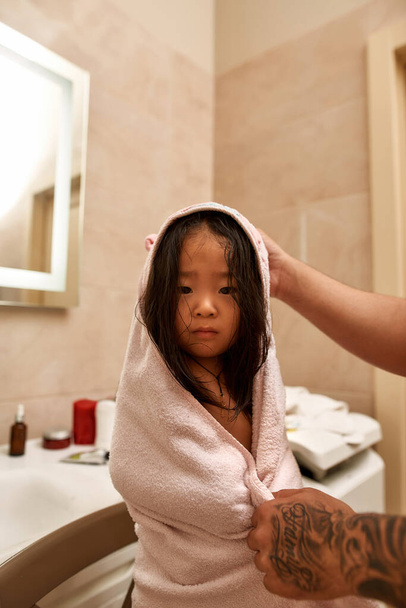 Частичный отец носит полотенце на мокрой азиатской маленькой дочери, смотрит в камеру в ванной. Мужчина и девушка проводят время вместе. Гигиена. Семейные отношения и забота. Родительство и отцовство. Доброе утро - Фото, изображение