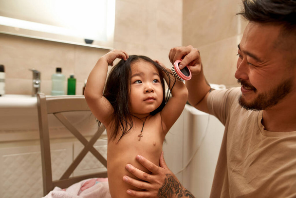 Smiling asian father combing hair of little daughter with comb in bathroom. Dark haired girl with naked torso. Hair care. Family relationship and caring. Parenting and fatherhood. Morning time - Photo, Image