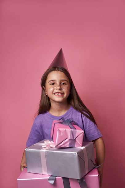 Smiling caucasian little girl with gift boxes look at camera. Kid wear party hat with colorful sequins on face. Holiday and event. Modern youngster lifestyle. Pink background. Studio shoot. Copy space - Photo, image