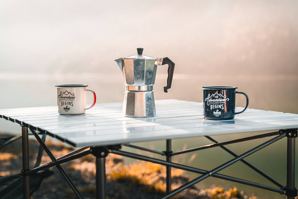 A classic moka pot and two matching coffee mugs sit on a sturdy folding table, ready for a morning pick-me-up while camping. Warm morning vibes for a relaxing coffee break in the great outdoors - Foto, Imagen