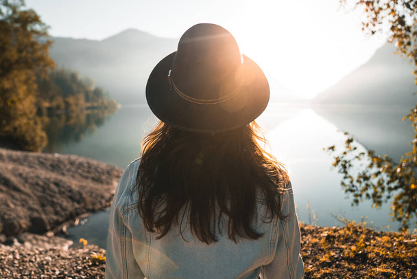 Вид сзади на модную молодую женщину с длинными волосами, стильную джинсовую куртку и гладкую черную шляпу, смотрящую на отражение озера и солнечный луч. Тихая вода, спокойная атмосфера, захватывающие дух пейзажи - Фото, изображение