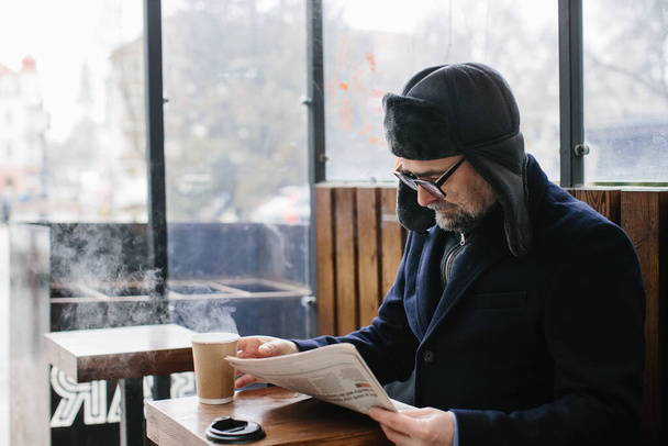 Un hombre de pelo gris con ropa de abrigo lee un periódico fresco y bebe té caliente o café en la calle. - Foto, Imagen