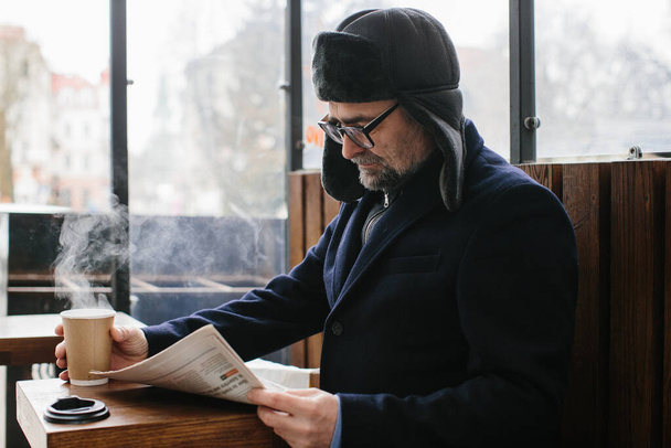 A gray-haired man in warm clothes reads a fresh newspaper and drinks hot tea or coffee on the street. - Foto, Imagem