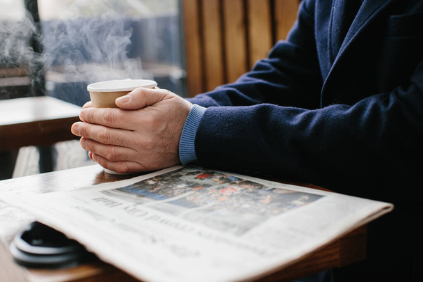 Egy meleg ruhás ősz hajú férfi friss újságot olvas, és forró teát vagy kávét iszik az utcán.. - Fotó, kép
