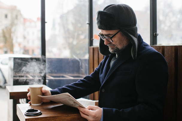 Šedovlasý muž v teplých šatech čte čerstvé noviny a pije horký čaj nebo kávu na ulici. - Fotografie, Obrázek