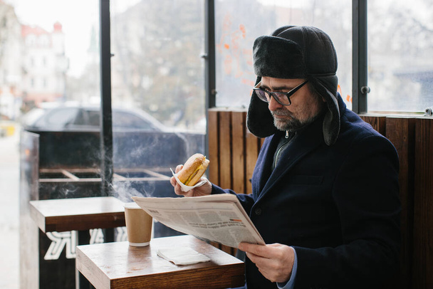 Vousatý muž středního věku jí chutný burger a čte dnešní noviny na městské ulici v zimě. Koncept pouličního jídla. - Fotografie, Obrázek