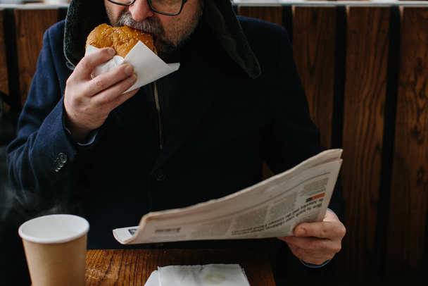 Ένας μεσήλικας γενειοφόρος τρώει ένα νόστιμο μπέργκερ και διαβάζει τη σημερινή εφημερίδα σε ένα δρόμο της πόλης το χειμώνα. Έννοια τροφίμων δρόμου. - Φωτογραφία, εικόνα